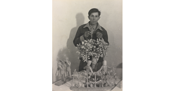 A black-and-white photograph of a medium-skinned man standing behind a wood sculpture of trees and human figures.