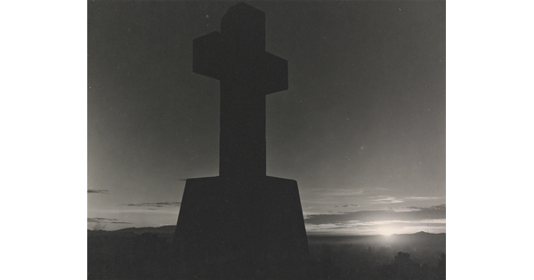A black-and-white photograph of a stone cross in silhouette and the sun peeking out from the horizon.