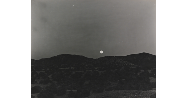 A black-and-white photograph of the moon rising above a mountain landscape.