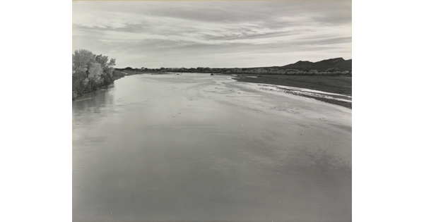 A black-and-white photograph of a calm river with trees on the left shore and mountains in the distance.