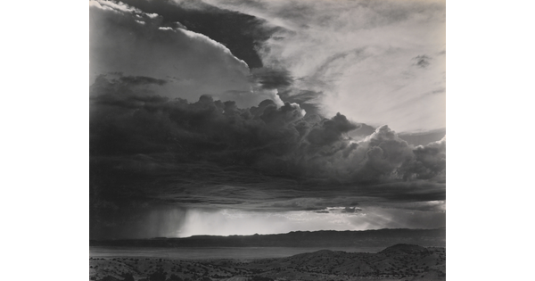 A black-and-white photograph of torrential rain and dark, heavy storm clouds passing into a brightly lit plain.