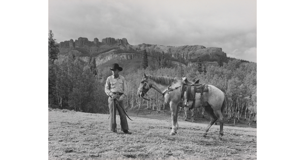 A black-and-white photograph of a White cowboy holding the reins of a horse in front of mountains.