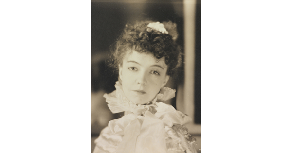 A black-and-white portrait photograph of a White woman with curly hair pulled on top of her head, wearing a top with a ruffled collar.