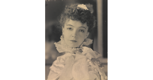 A black-and-white portrait photograph of a White woman with curly hair pulled on top of her head, wearing a top with a ruffled collar.