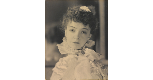 A black-and-white portrait photograph of a White woman with curly hair pulled on top of her head, wearing a top with a ruffled collar.