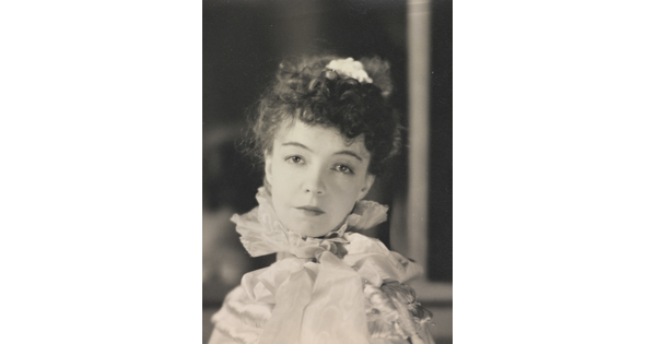 A black-and-white portrait photograph of a White woman with curly hair pulled on top of her head, wearing a top with a ruffled collar.