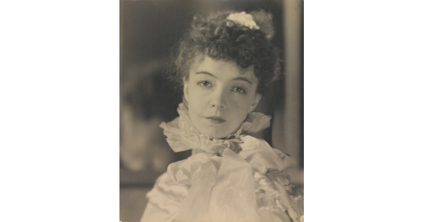 A black-and-white portrait photograph of a White woman with curly hair pulled on top of her head, wearing a top with a ruffled collar.