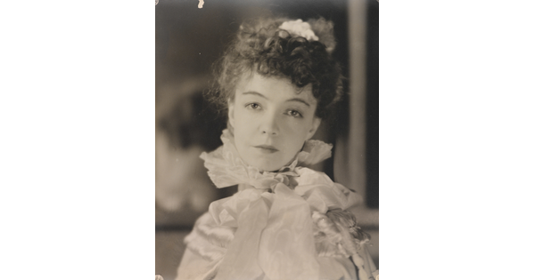 A black-and-white portrait photograph of a White woman with curly hair pulled on top of her head, wearing a top with a ruffled collar.