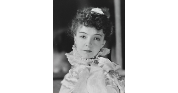 A black-and-white portrait photograph of a White woman with curly hair pulled on top of her head, wearing a top with a ruffled collar.