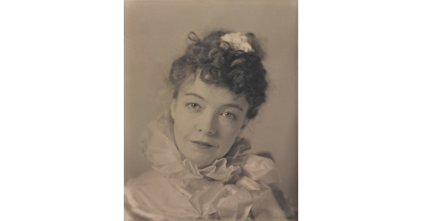 A black-and-white portrait photograph of a White woman with curly hair pulled on top of her head, wearing a top with a ruffled collar.