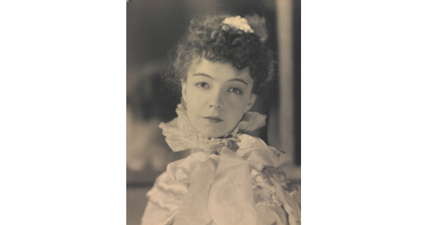 A black-and-white portrait photograph of a White woman with curly hair pulled on top of her head, wearing a top with a ruffled collar.