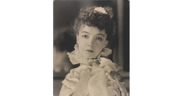 A black-and-white portrait photograph of a White woman with curly hair pulled on top of her head, wearing a top with a ruffled collar.