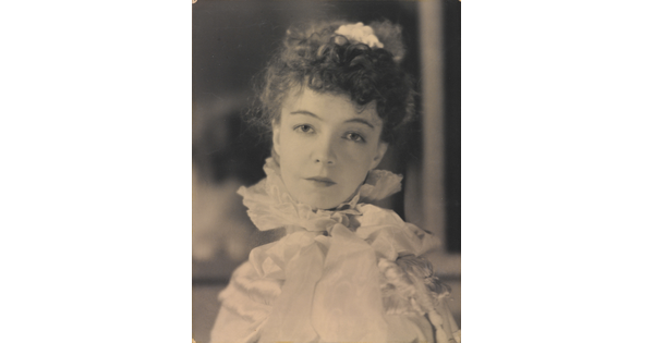 A black-and-white portrait photograph of a White woman with curly hair pulled on top of her head, wearing a top with a ruffled collar.
