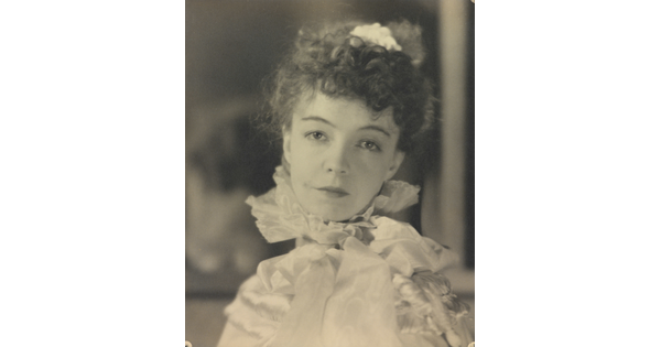 A black-and-white portrait photograph of a White woman with curly hair pulled on top of her head, wearing a top with a ruffled collar.