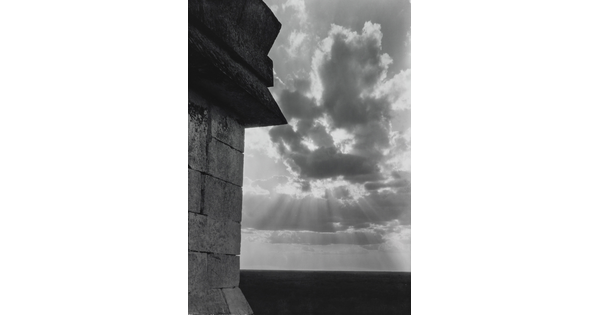 A black-and-white photograph of the sun's rays bursting from clouds and a stark brick architectural feature in the left foreground.