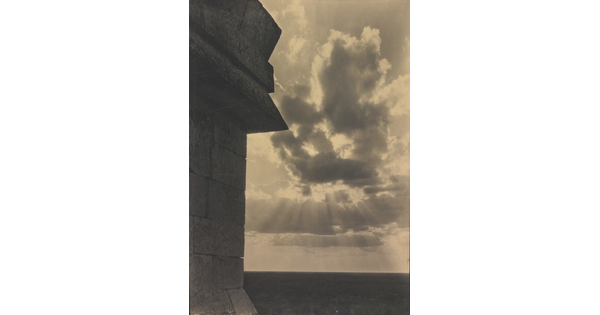 A sepia-toned photograph of the sun's rays bursting from clouds and a stark brick architectural feature in the left foreground.