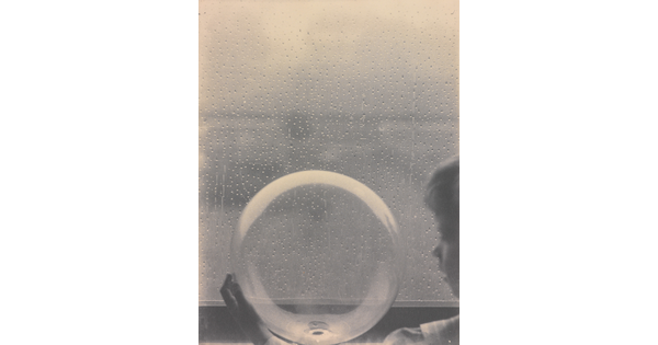 A black-and-white photograph of a White child looking at a clear glass globe in front of a rain-spattered window.