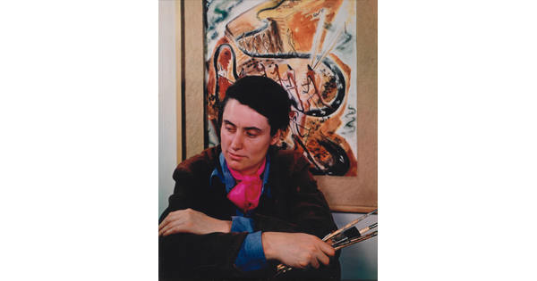 A color portrait photograph of a seated White woman holding paintbrushes in front of an abstract painting.