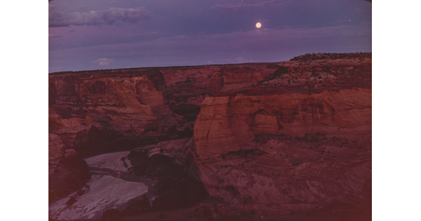 A color photograph of a full moon shining in a purple and blue sky over a steep, red, rocky canyon.