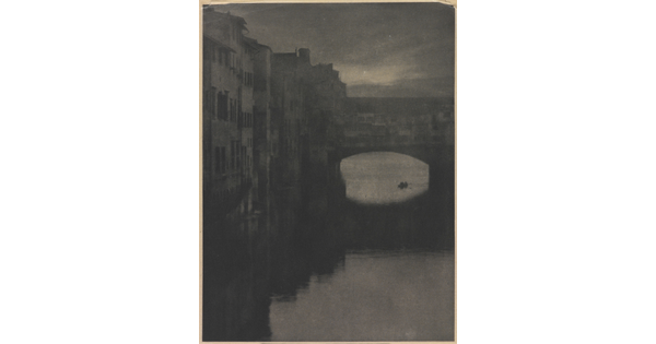 A sepia-toned photograph of a wall of tall buildings lining a river and an enclosed arched bridge stretching over the water.