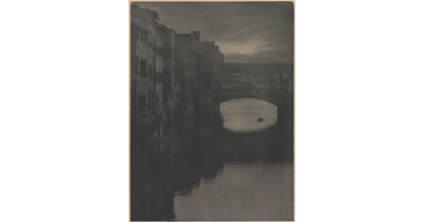 A sepia-toned photograph of a wall of tall buildings lining a river and an enclosed arched bridge stretching over the water.