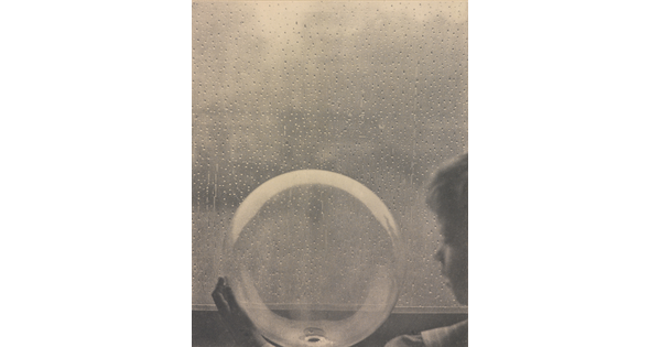 A black-and-white photograph of a White child looking at a clear glass globe in front of a rain-spattered window.