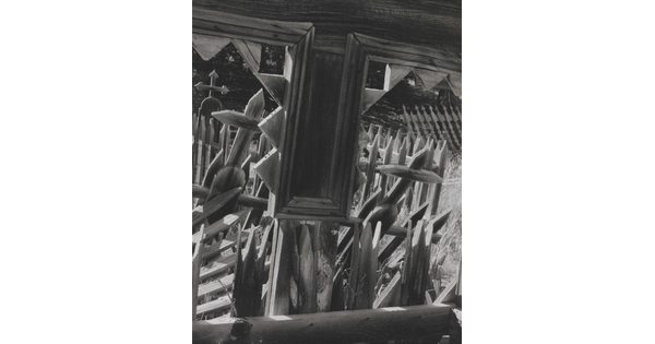 A black-and-white photograph looking through the windows of a wooden structure onto a cemetery with wooden crosses and fence posts.