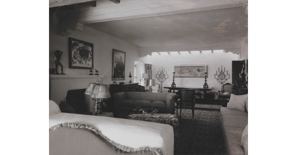 A black-and-white photograph of a formal living room in an adobe building with a fireplace, multiple couches, paintings, and sculptures.
