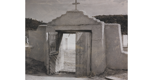 A black-and-white photograph of an adobe wall with a doorway that leads to a courtyard; a cross on the wall above the wooden doors.