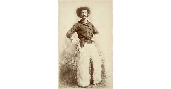 A sepia-toned formal portrait photograph of a cowboy posing in western wear, including white furry chaps, with his hands on his hips.