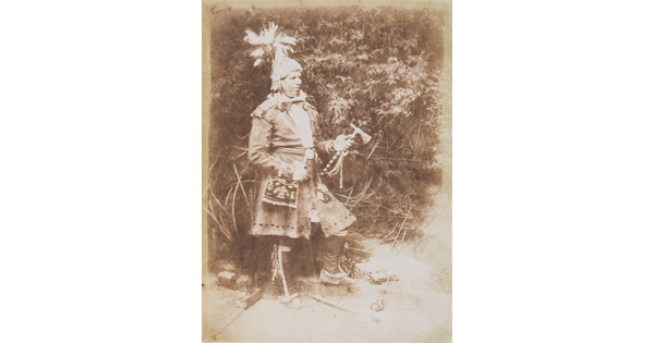 A sepia-toned photograph of an Indigenous man wearing traditional clothing and holding a tomahawk.