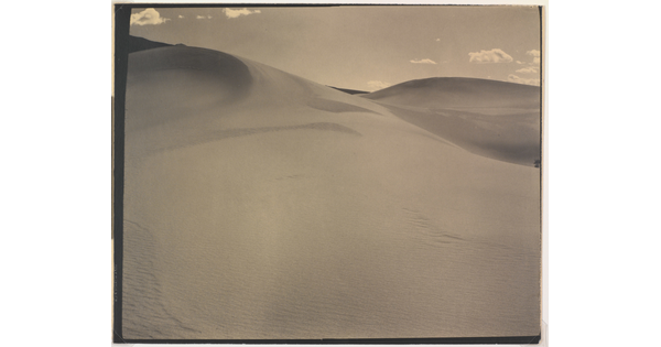 A sepia-toned photograph of a rippling sand dune rising up into a sky dotted with a few small clouds.
