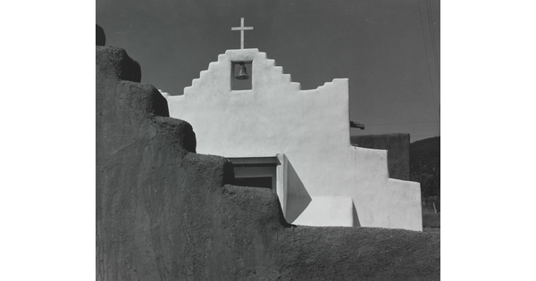 A black-and-white photograph of a terraced adobe bell tower with a cross on top, partially hidden by a terraced adobe wall.