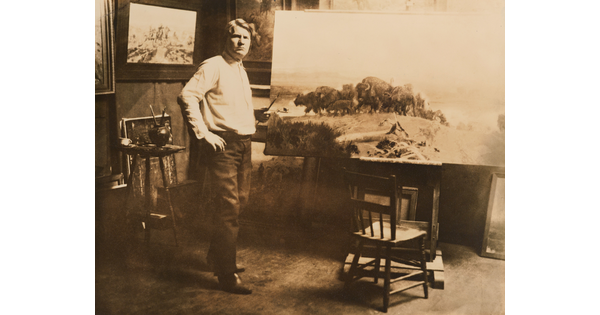 A sepia-toned photograph of a White man posing next to a large canvas that depicts a herd of bison.