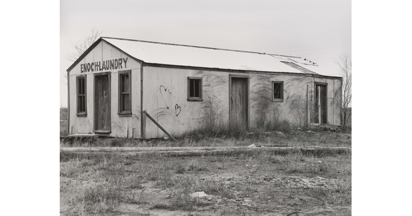 A black-and-white photograph of a one-story building with boarded up windows and graffiti on the walls; lettering above the door reads, "Enoch Laundry."