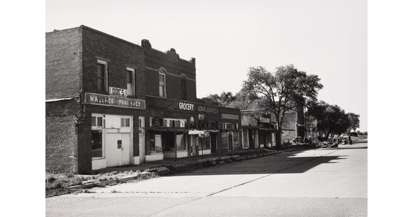 A black-and-white photograph of a paved street lined with brick buildings, some boarded up, casting stark shadows on the ground.