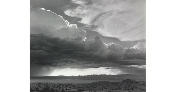 A black-and-white photograph of torrential rain and dark, heavy storm clouds passing into a brightly lit plain.