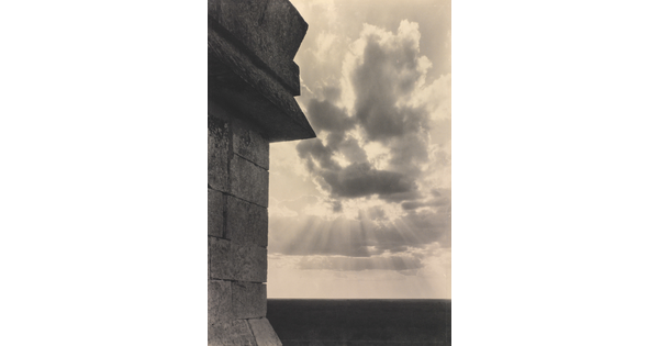A sepia-toned photograph of the sun's rays bursting from clouds and a stark brick architectural feature in the left foreground.