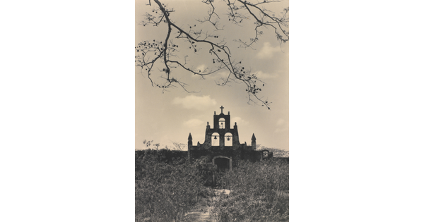A black-and-white photograph of the facade and terraced bell tower, topped with a cross, of an adobe church in a field of tall grass and weeds.