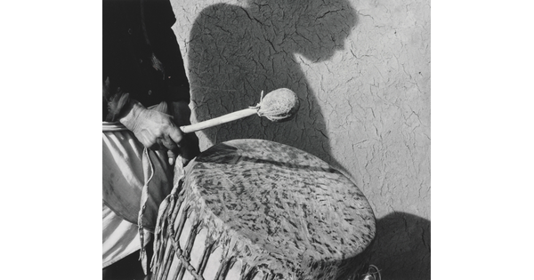 A black-and-white photograph of a person holding a drumstick over a traditional drum and the person's shadow in profile on the wall in the background.