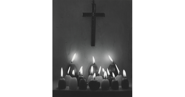 A black-and-white photograph of three rows of lit votive candles in front of a cross hanging on a wall.