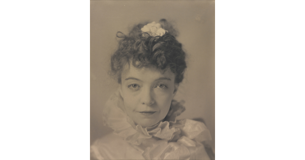 A black-and-white portrait photograph of a White woman with curly hair pulled on top of her head, wearing a top with a ruffled collar.
