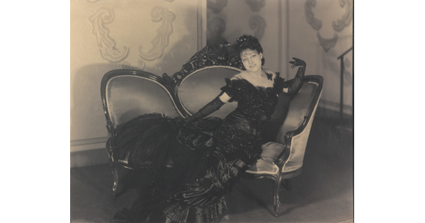 A black-and-white photograph of a White woman in a long, formal gown and elbow-length gloves posing on a sofa.