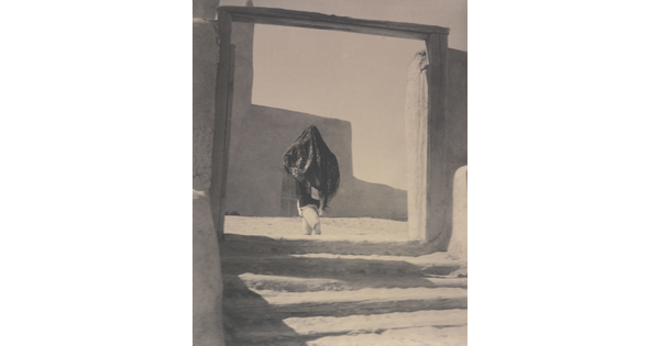A black-and-white photograph of a person wearing a long shawl over their head and torso while walking through the wood gate of an adobe building.