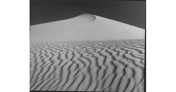 A sepia-toned photograph of a deeply-rippled sand dune rising to a point into the sky.