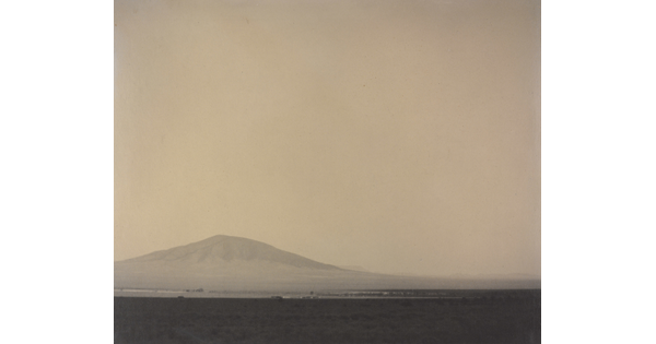 A black-and-white photograph of a flat, desert landscape with a mountain rising up in the hazy distance.