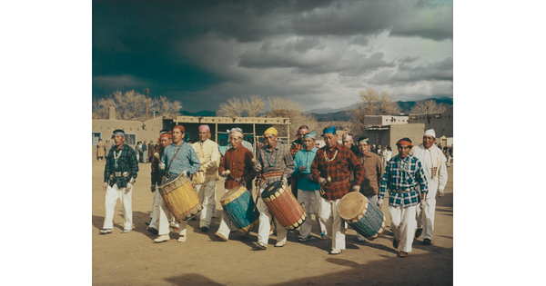 A color photograph of Indigenous people walking, four of them carrying tall drums.