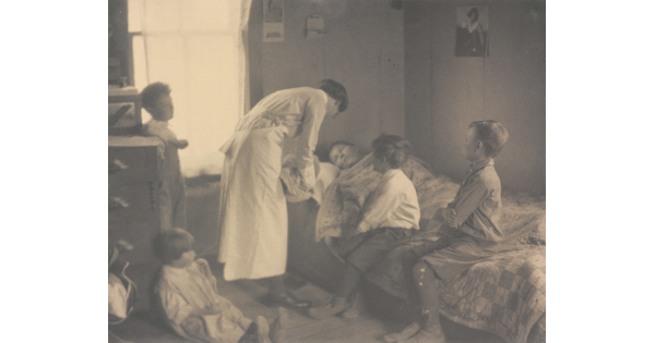 A black-and-white photograph of a nurse bending over a person in a bed, two children sitting on the bed, and two children behind the nurse.