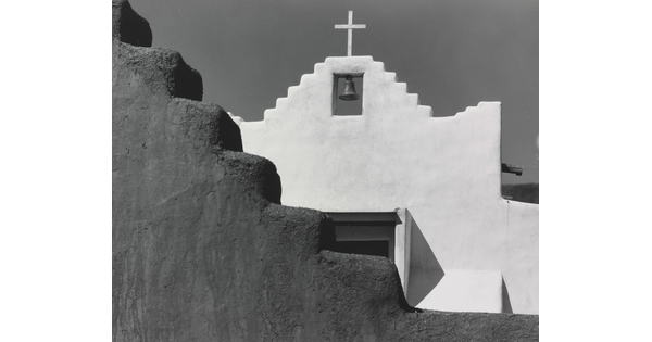 A black-and-white photograph of a terraced adobe bell tower with a cross on top, partially hidden by a terraced adobe wall.