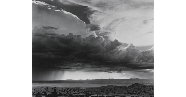 A black-and-white photograph of torrential rain and heavy storm clouds passing into a brightly lit plain.